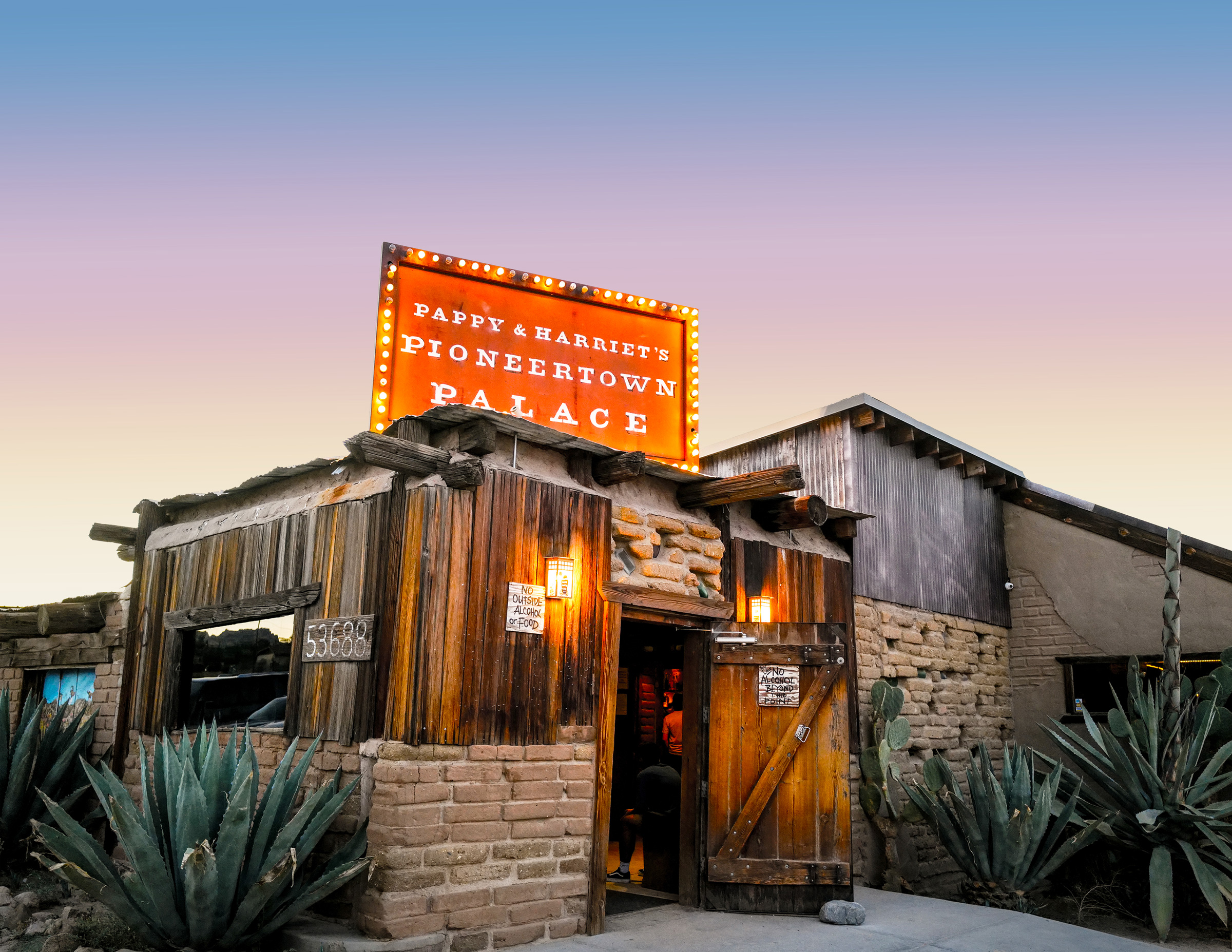 Pappy & Harriet’s Pioneertown Palace at dusk, with its illuminated orange sign above a rustic wood-and-stone entrance framed by agaves.