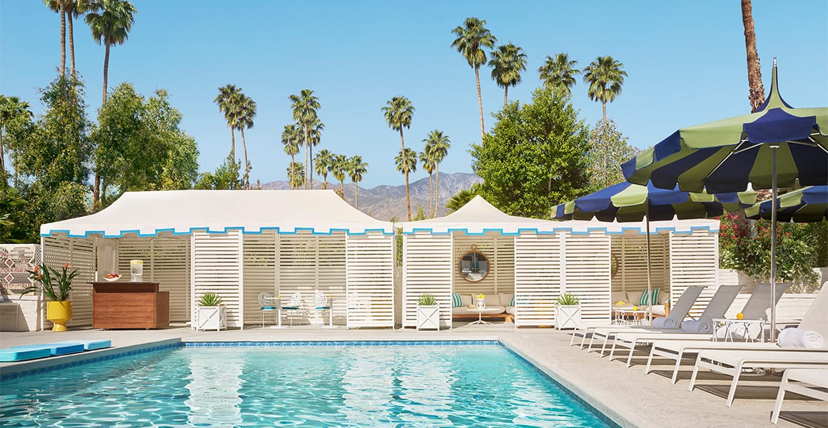 Turquoise pool at Parker Palm Springs with white cabanas, striped parasols, palm trees, and desert mountains.