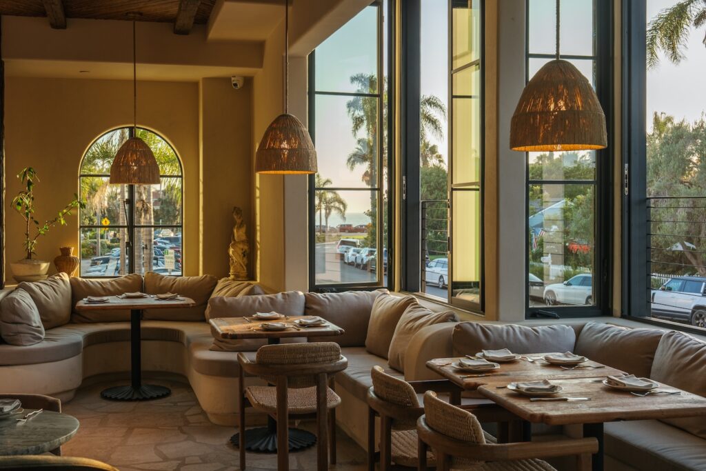 Window-side dining room at By The Sea in Coronado, with curved upholstered seating and woven pendant lights.