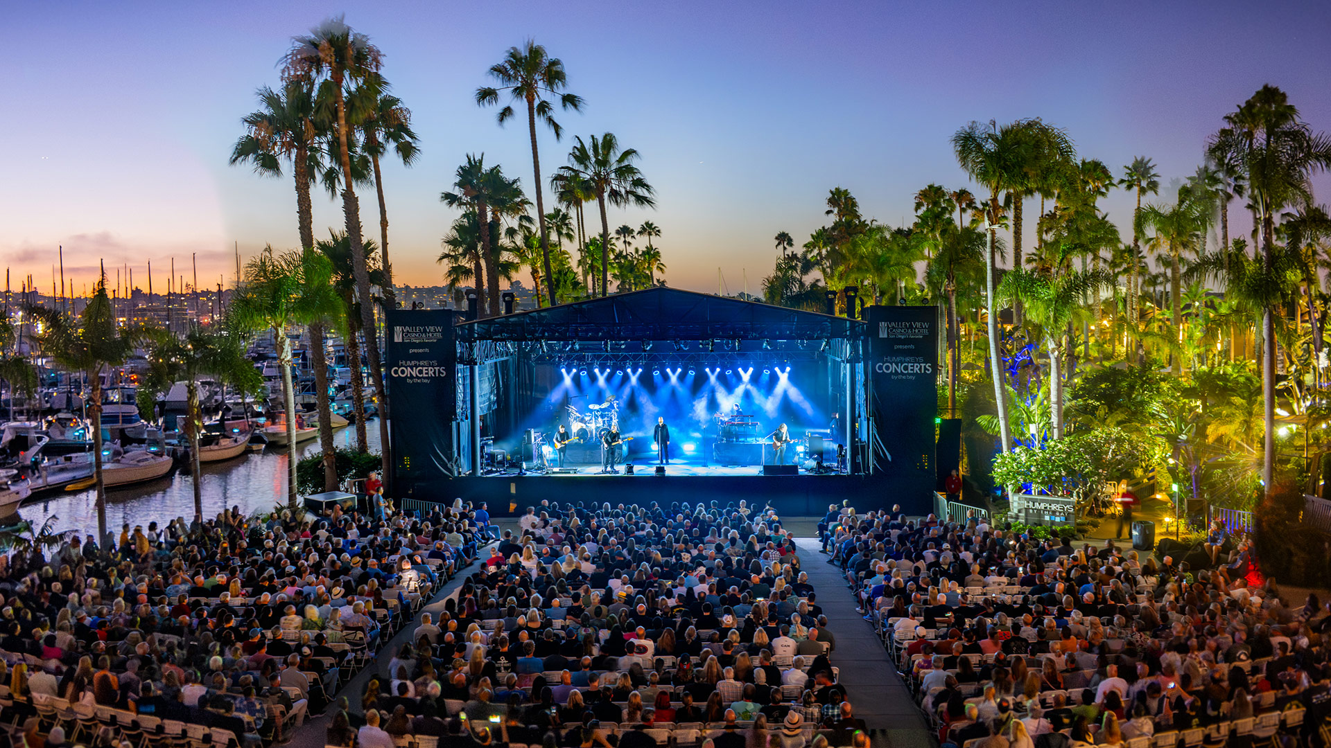 A concert audience faces a blue-lit waterfront stage surrounded by palm trees and boats at Humphreys Concerts by the Bay.