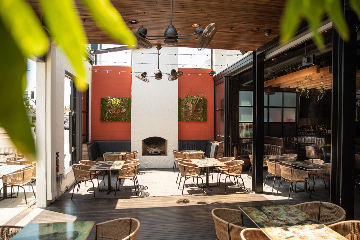 La Puerta Mission Hills patio with woven chairs, wooden tables, and a white fireplace against a terracotta-colored wall.