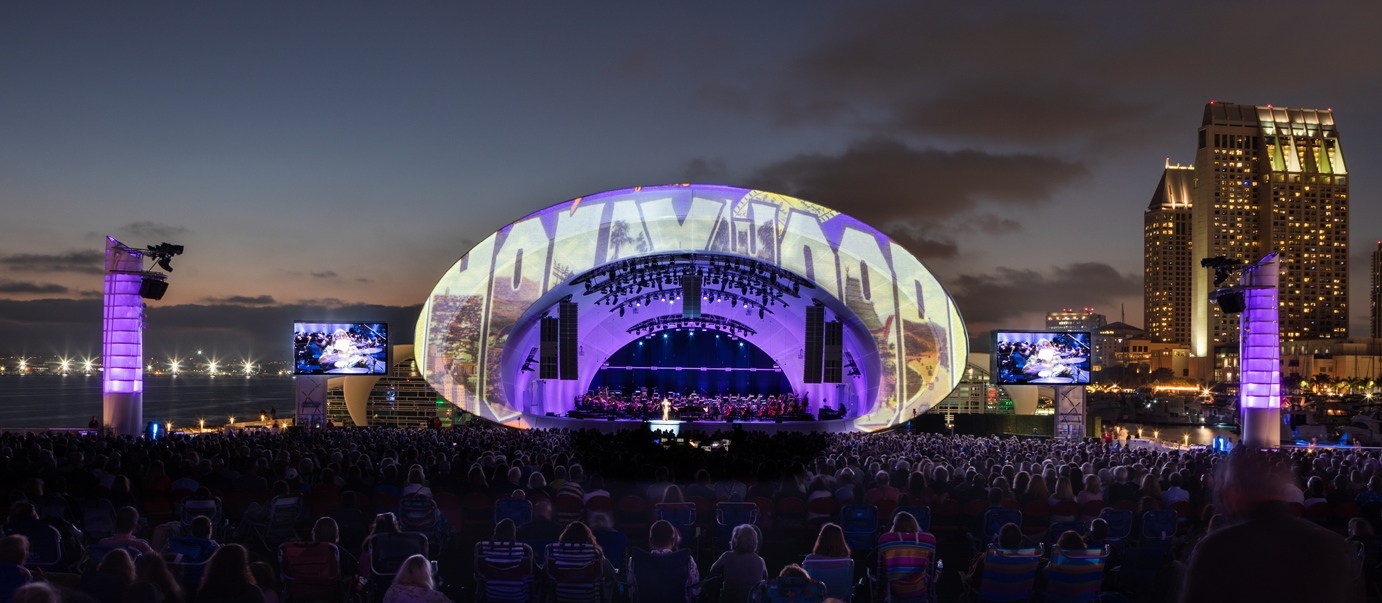 Audience watches an orchestra beneath the illuminated Rady Shell at dusk beside San Diego Bay.